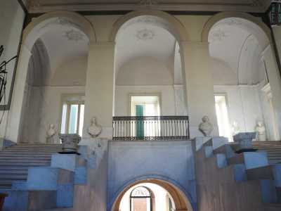 Percorriamo la scalinata in marmo ornata da mezzi-busto e capitelli giungendo infine al primo livello. Tre arcate simmetriche, sorrette da robusti pilastri, inaugurano il piano superiore chiuso da volte a botte...